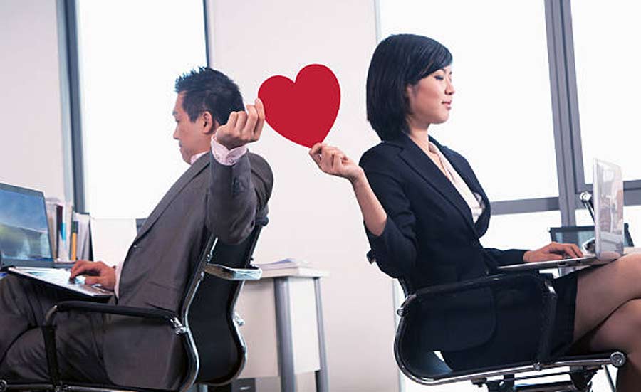 Office scene with a woman holding a heart sign behind a man.